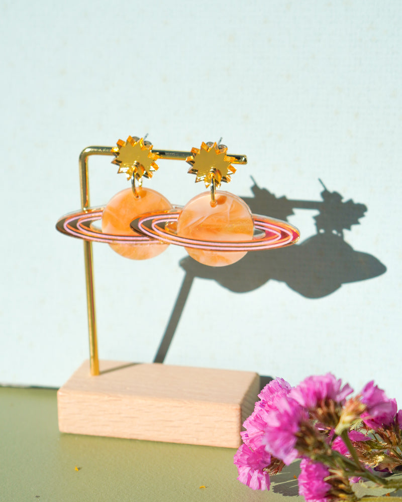 Flowature's Saturn Earrings with a blue backdrop and pink flowers in the foreground.