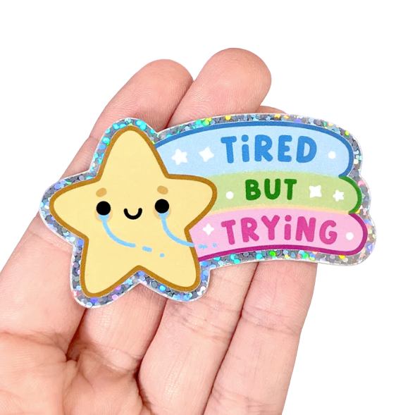 A tiny crying star sticker with a rainbow in it's wake reading: Tired But Trying