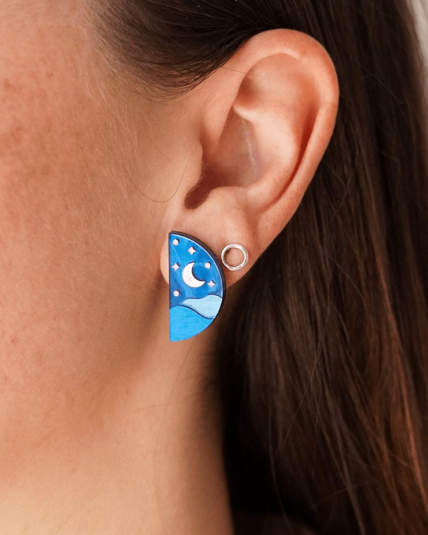 Flowatures 'Moon' stud. The moon is a bright silver with blue waves below and silver stars surrounding it.