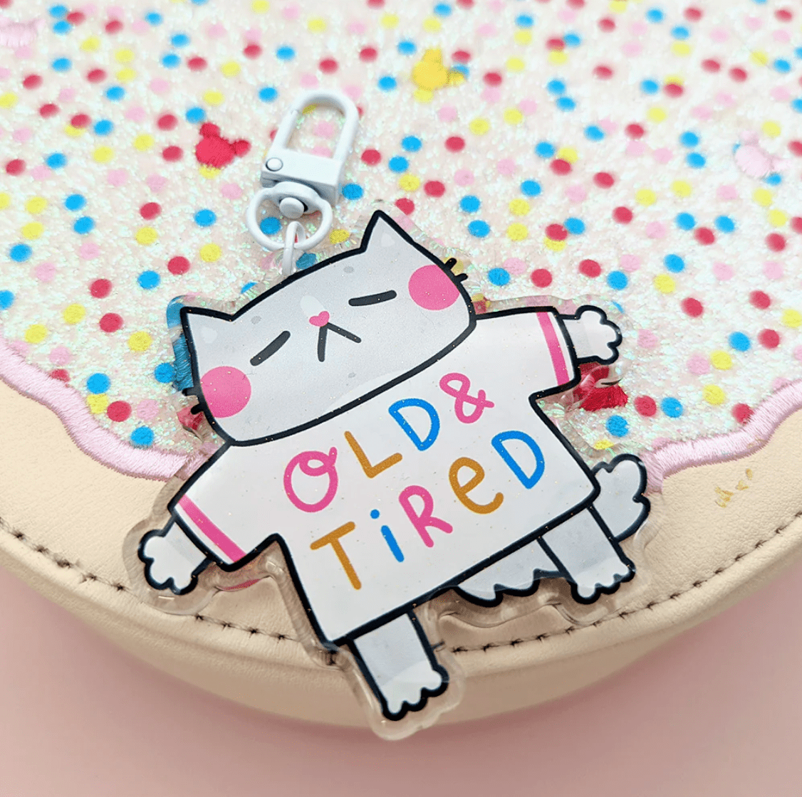 A super cute keychain by Fox & Cactus: a sleeping cat with pink cheeks and a heart note with a tee shirt that reads "Old & Tired" on the front and "Go Away" on the back.