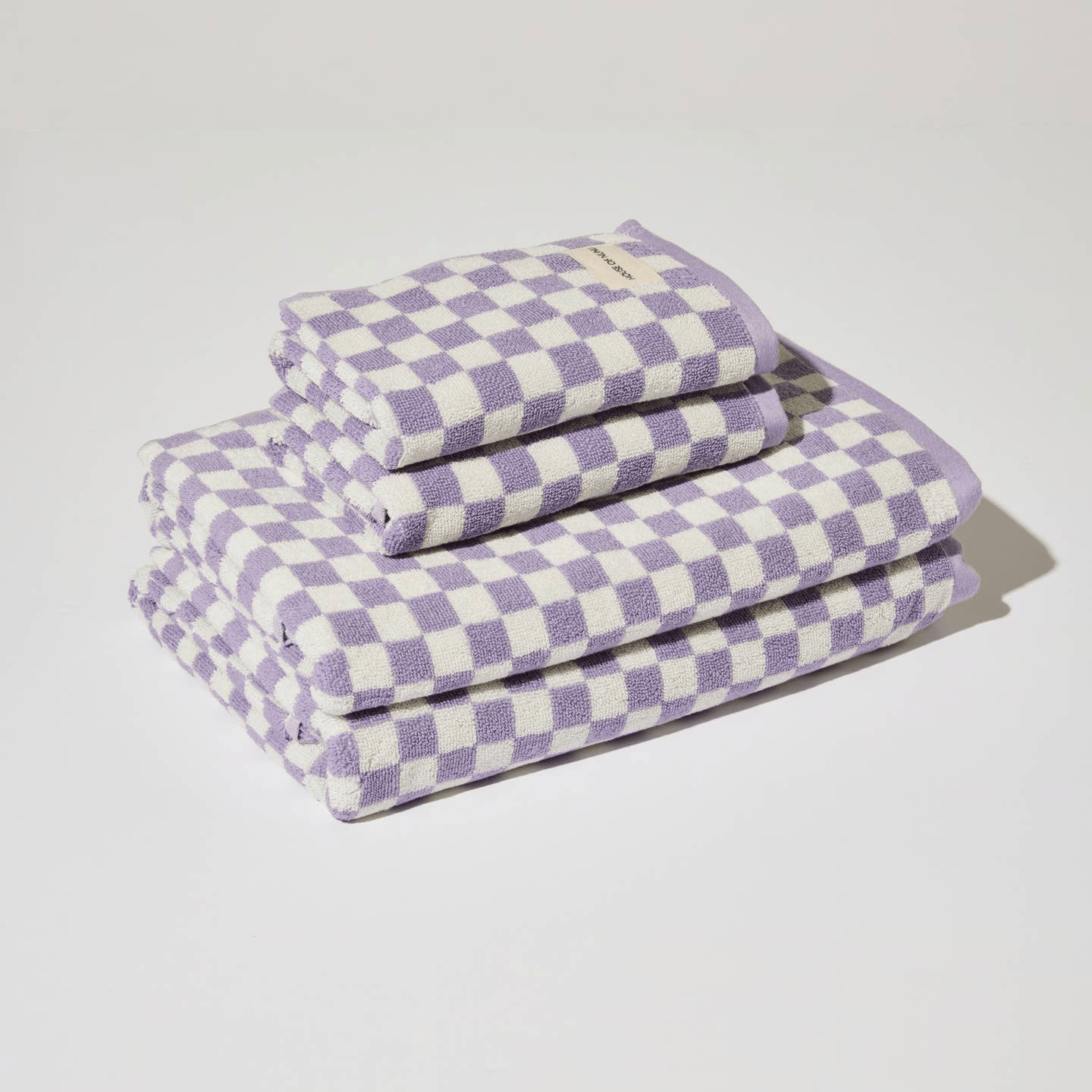 A cute bath towel pile by House of Nunu. Two hand towels and 2 bath towels are folded into perfect rectangles on top of each other. The print features lilac checks on a white background.