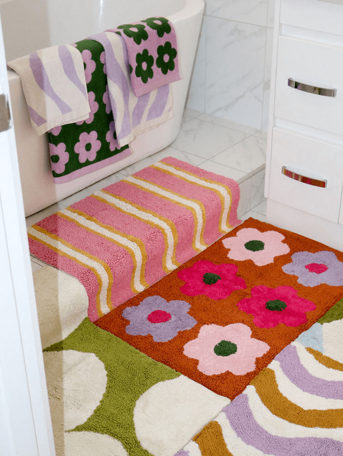 A pink and mustard yellow stripe bath mat by Mosey Me next to other colourful bath mats on a bathroom floor.