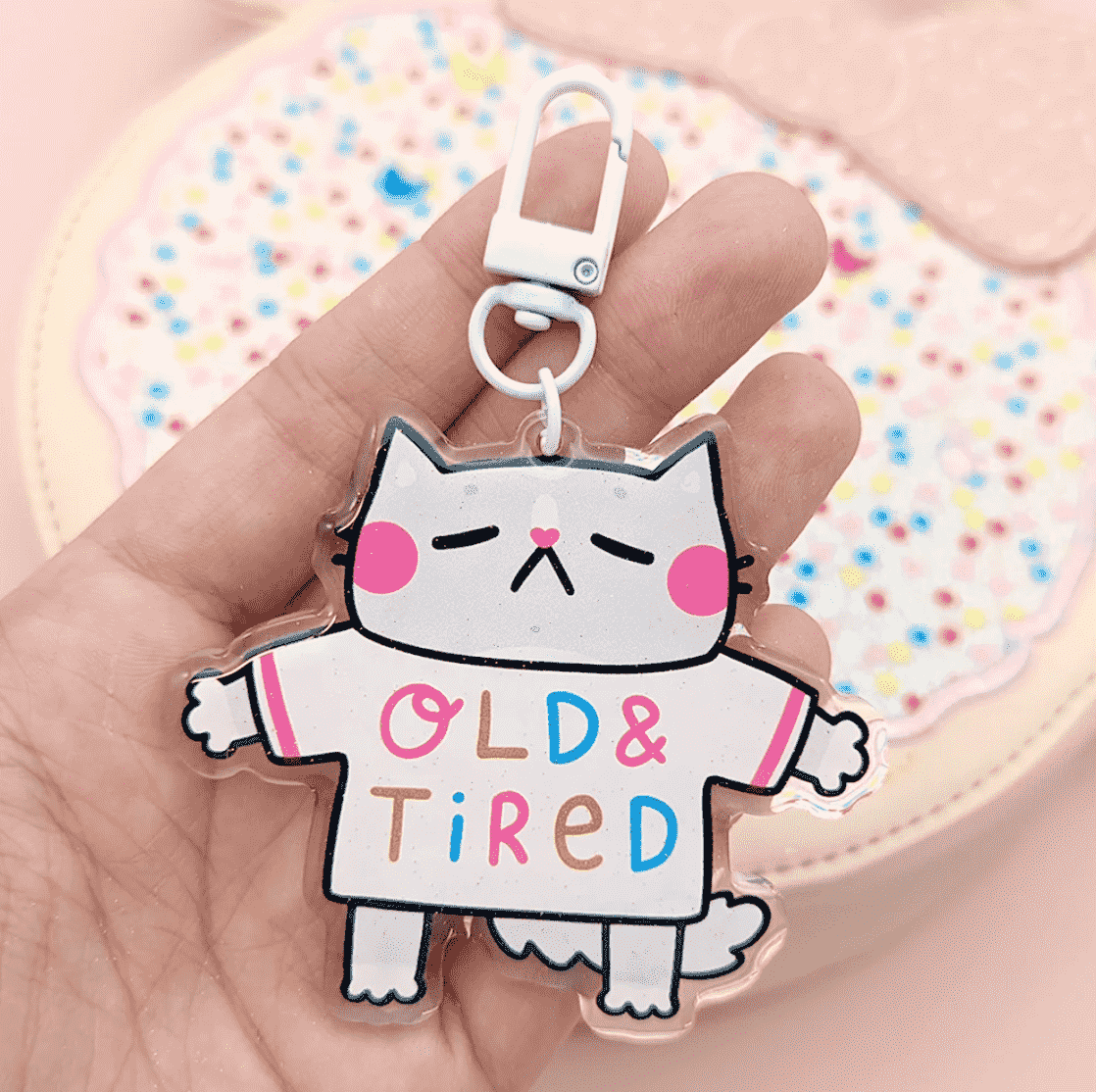 A super cute keychain by Fox & Cactus: a sleeping cat with pink cheeks and a heart note with a tee shirt that reads "Old & Tired" on the front and "Go Away" on the back.
