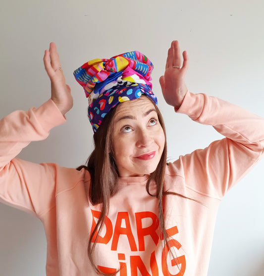Carly Spiteri wearing a peach sweater reading "DARLING" in capital letters & a bright red font. She's wearing a stack of her favourite, colourful headbands on her head. 