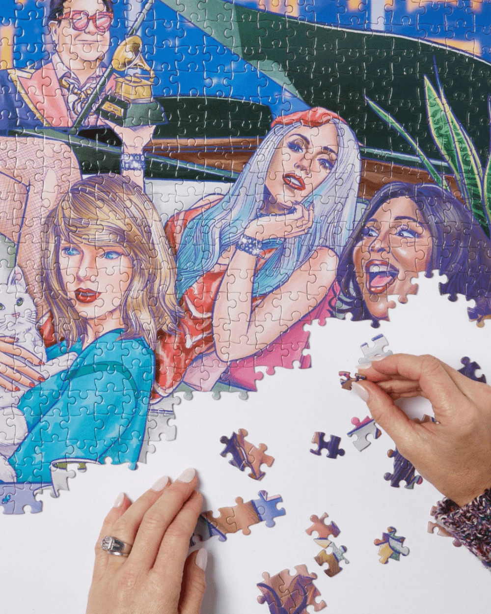 Half of the puzzle as a work in progress with hands finishing the piece. This part features Elton John, Lizzo, Lady Gaga and Taylor Swift with her cat. 