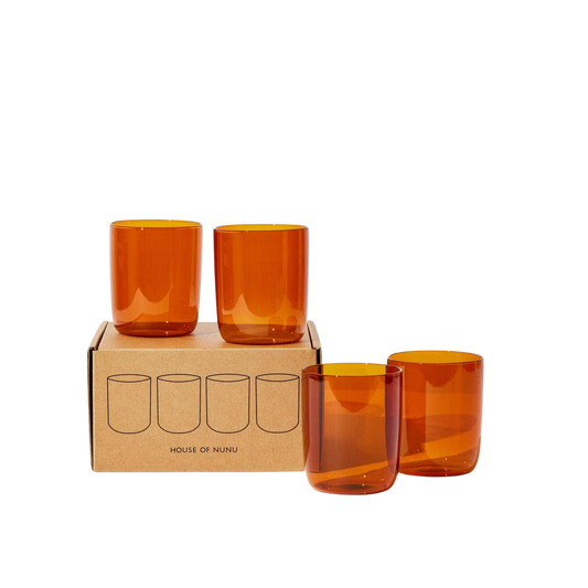 House of Nunu Amber Glass Set: Four amber coloured drinking glasses stacked on top of their brown box in front of a white backdrop. 