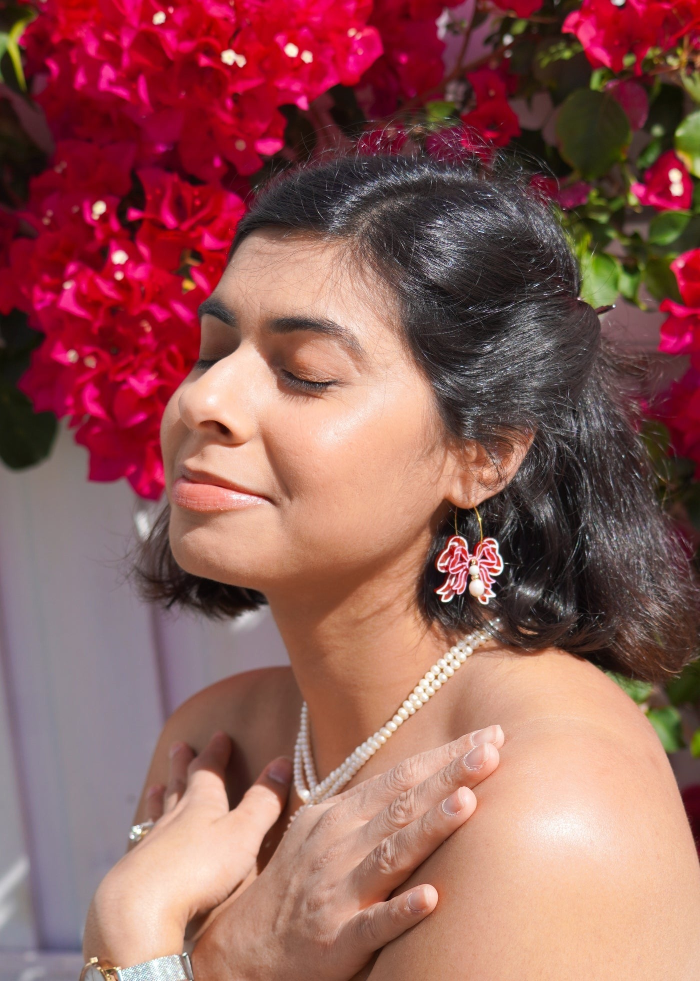 A flowature model wearing Bow Blossom Earrings in front of bright red flowers. 