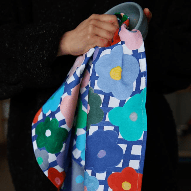 Shuh Lee's colourful Flower Tea Towel, adorned with bright multicoloured flowers being used on a pastel blue bowl. 