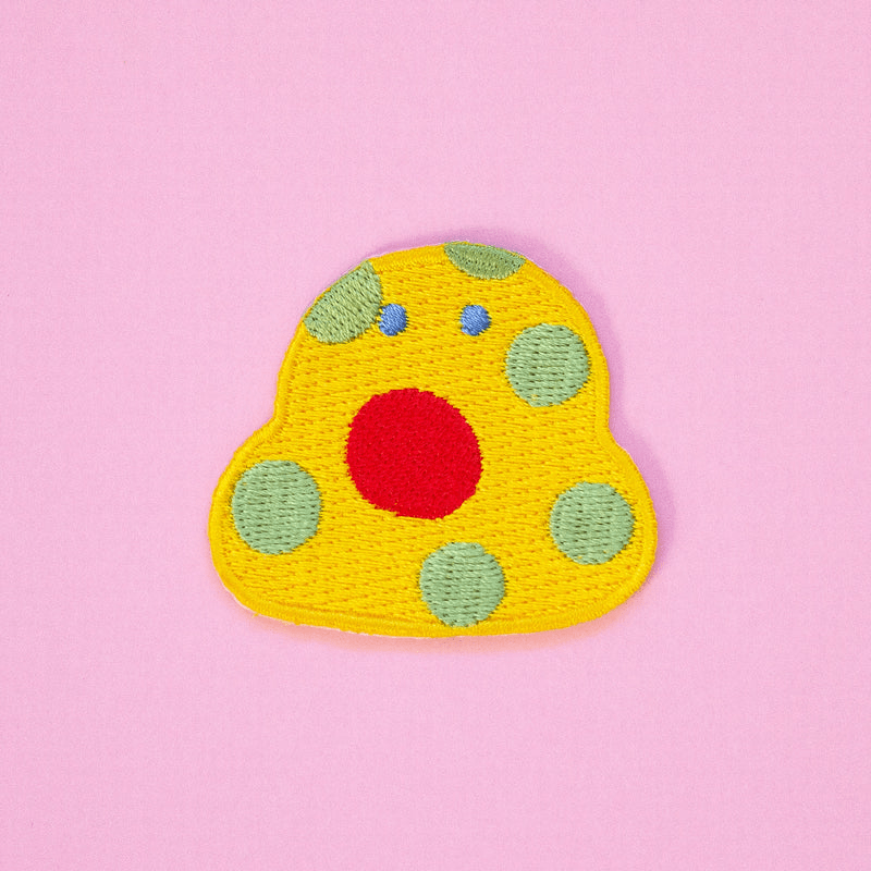 Shuh Embroidery Patch: A quirky little yellow blob with pastel green spots, tiny blue eyes and a large circular red mouth. 