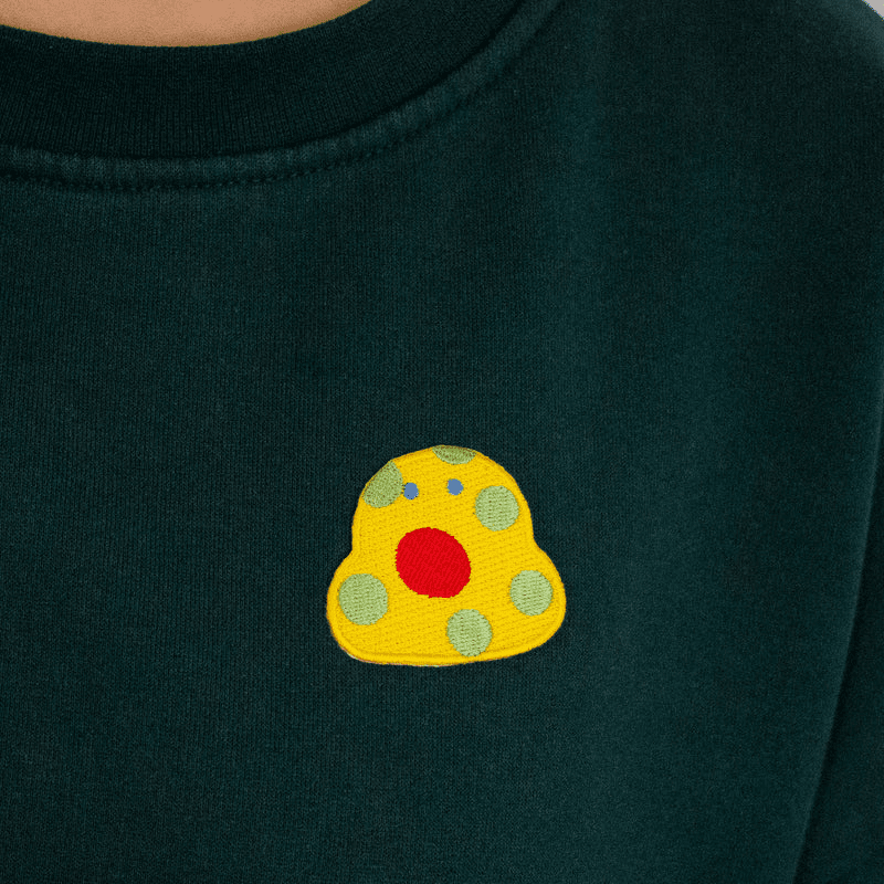 Shuh Embroidery Patch: A quirky little yellow blob with pastel green spots, tiny blue eyes and a large circular red mouth. 