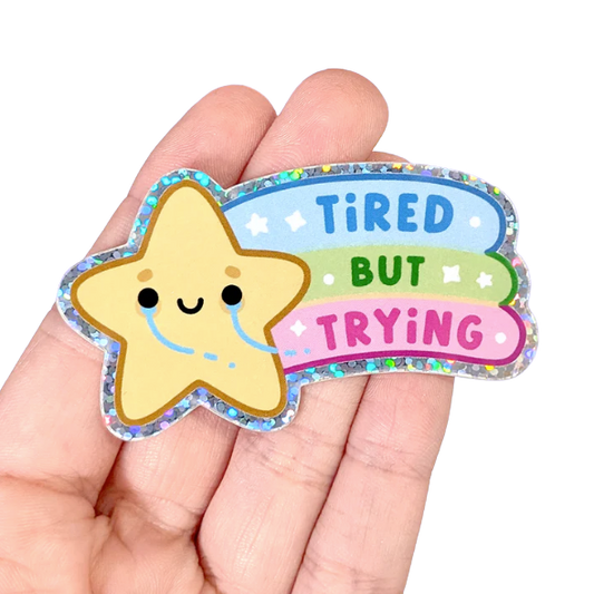 A tiny crying star sticker with a rainbow in it's wake reading: Tired But Trying