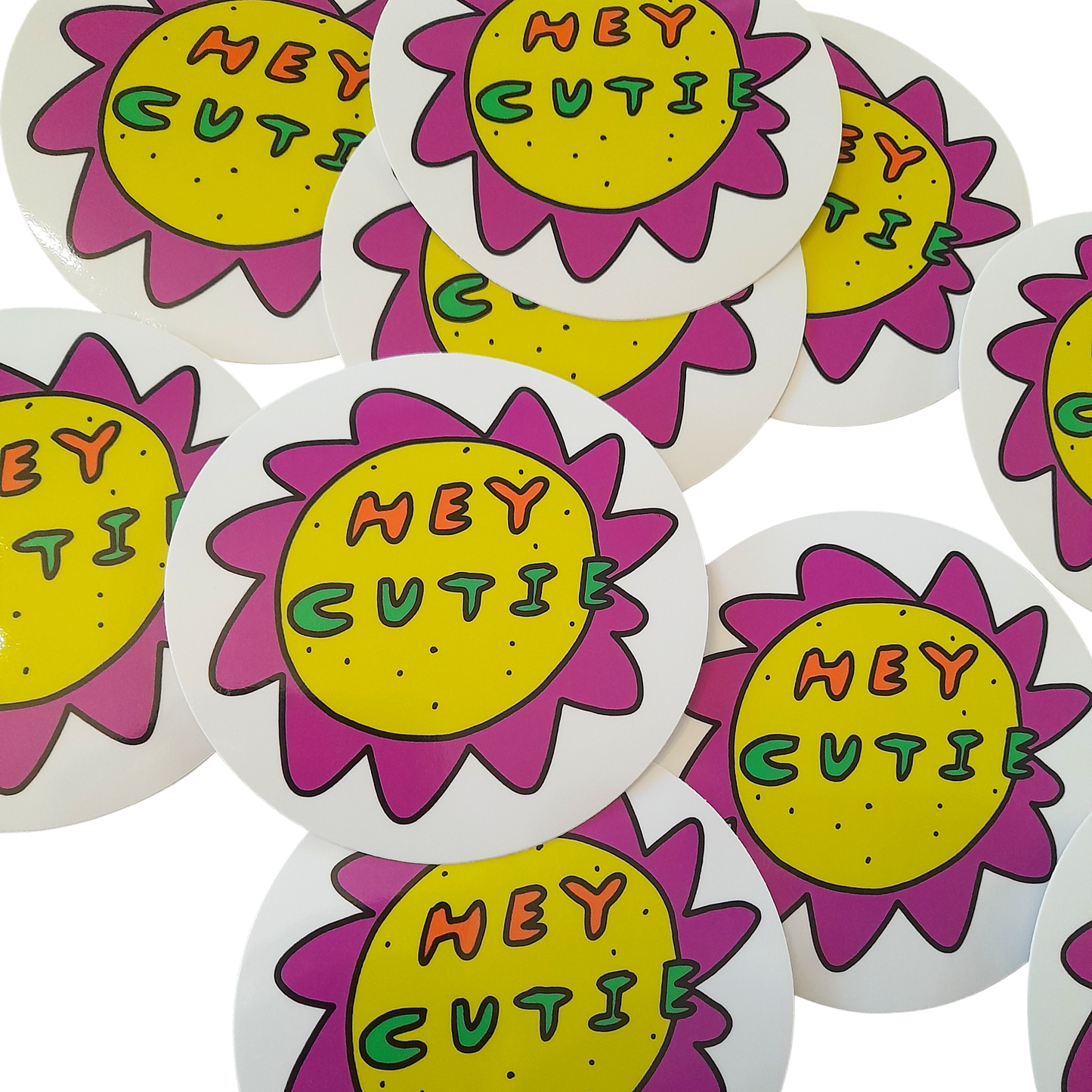 Oh Hey Cutie Sticker by Tegan Iversen is a circle sticker with a white background. It features a playful, quirky flower with a yellow centre and purple peak like petals around it. Inside it reads HEY in orange and CUTIE in green. 