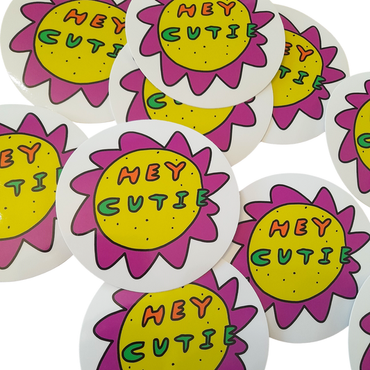 Oh Hey Cutie Sticker by Tegan Iversen is a circle sticker with a white background. It features a playful, quirky flower with a yellow centre and purple peak like petals around it. Inside it reads HEY in orange and CUTIE in green. 