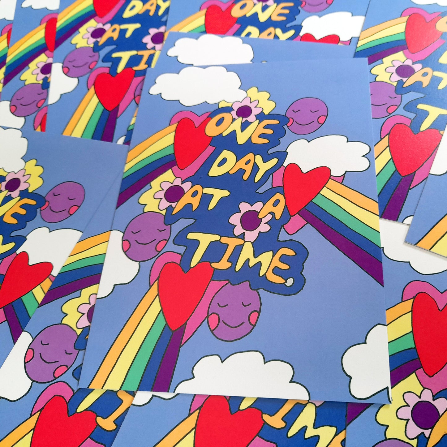 "One Day at a Time" postcard print by Tegan Iversen, illustrated in bold retro style with a rainbow, red hearts, fluffy white clouds and a smiling purple face on a blue background, with the words "One Day At A Time" in yellow lettering.