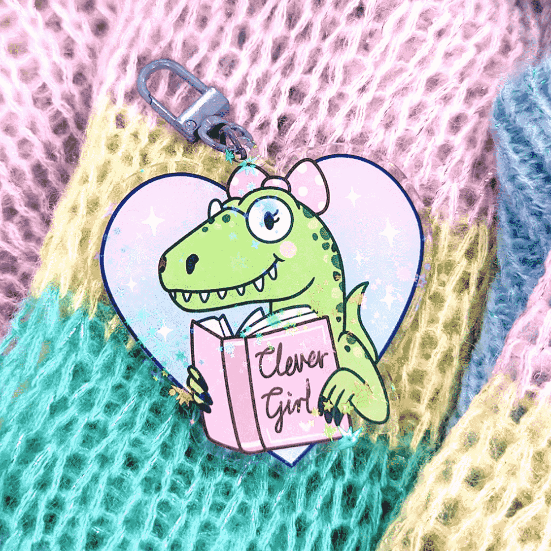 A cute keychain by Fox & Cactus of a happy green dinosaur. They have round glasses on, pink cheeks, a toothy grin and a pink bow in their hair. They have a book that reads "Clever Girl" 