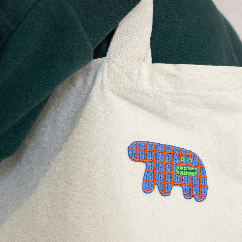 Shuh Lee Embroidery Patch: A funny abstract, surreal animal. It has four short  legs with a thick, short train. It's body is a sea blue with patchwork red lines across. It has a funny, open mouth showing its green teeth with small green eyes in the same shade. 