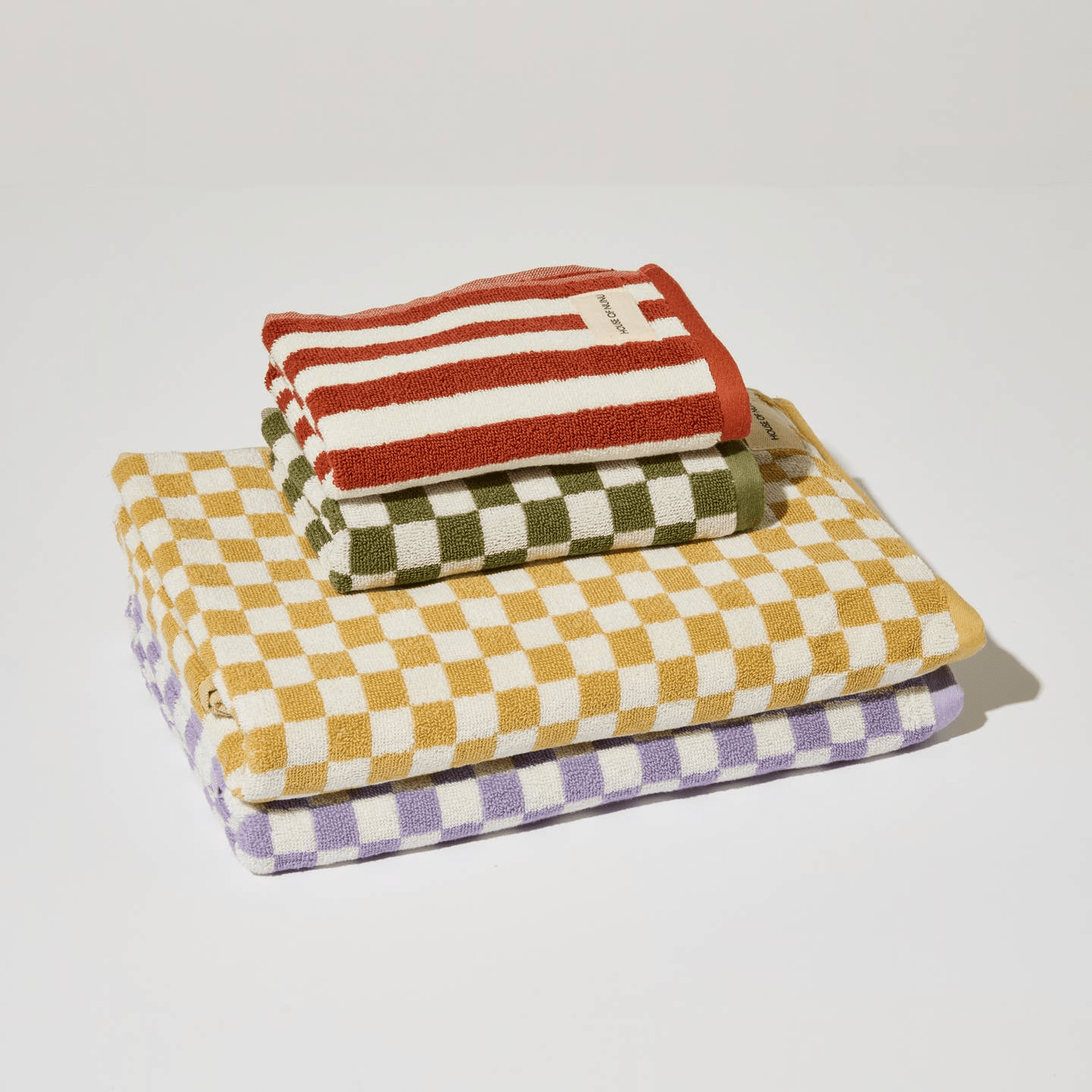 A cute pile of colourful towels by House of Nunu. Featuring two hand towels in a red stripe and green check, with two towels in mustard yellow and lilac underneath. 
