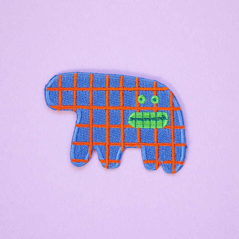 Shuh Lee Embroidery Patch: A funny abstract, surreal animal. It has four short  legs with a thick, short train. It's body is a sea blue with patchwork red lines across. It has a funny, open mouth showing its green teeth with small green eyes in the same shade. 